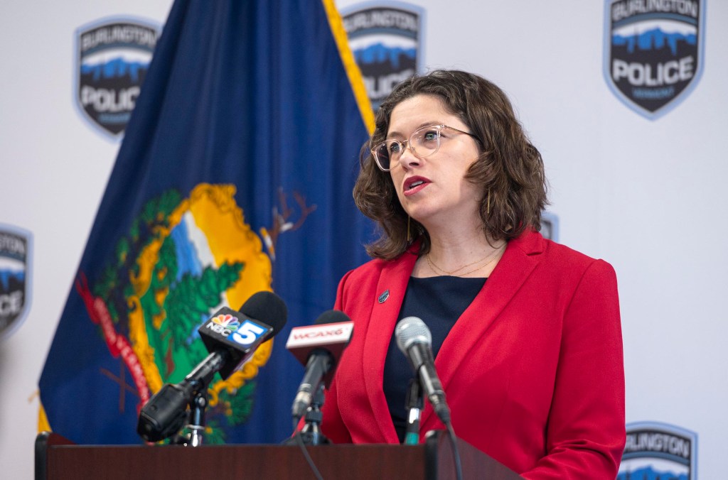 A person in a red blazer speaks into multiple microphones at a podium, with a flag and police logos in the background.