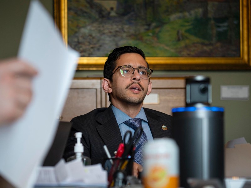 A man in a suit and glasses sits at a desk with office supplies, papers, a can, and a bottle of hand sanitizer, with a painting on the wall behind him.