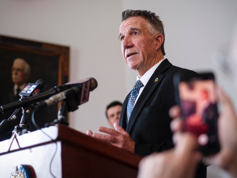 A man in a suit speaks at a podium with microphones, while another person takes a photo with a smartphone. A portrait hangs on the wall in the background.