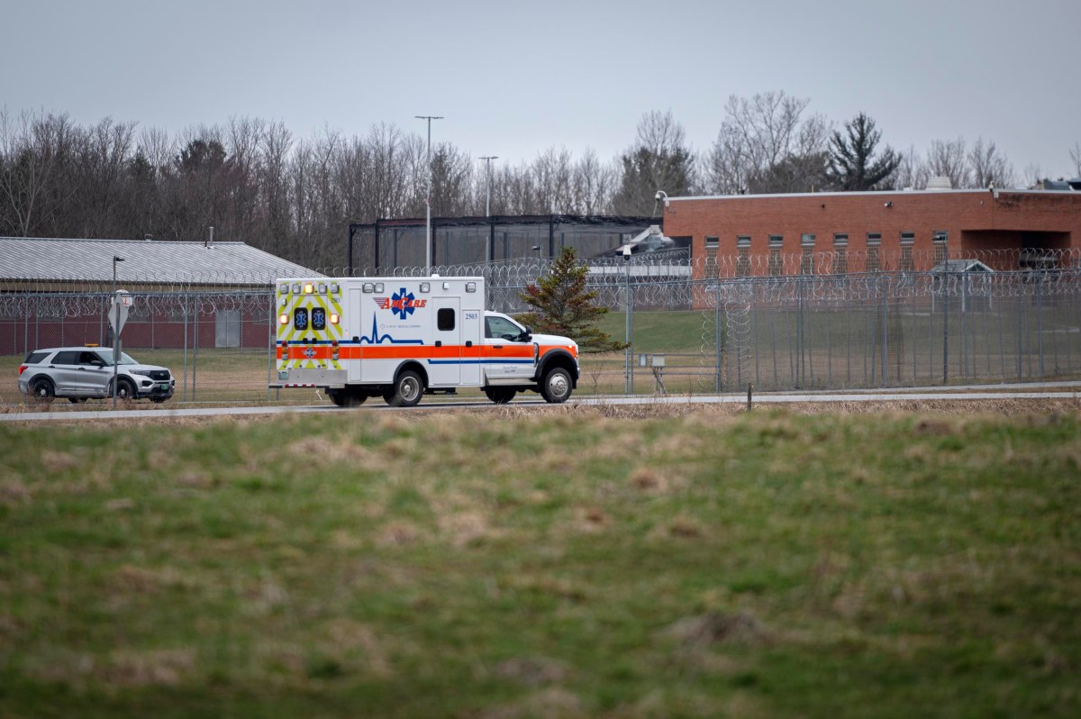 An ambulance drives past a barbed-wire fence surrounding a correctional facility, with a police SUV nearby and overcast sky overhead.