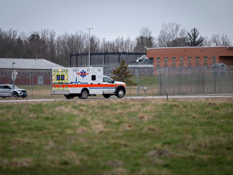 An ambulance drives past a barbed-wire fence surrounding a correctional facility, with a police SUV nearby and overcast sky overhead.