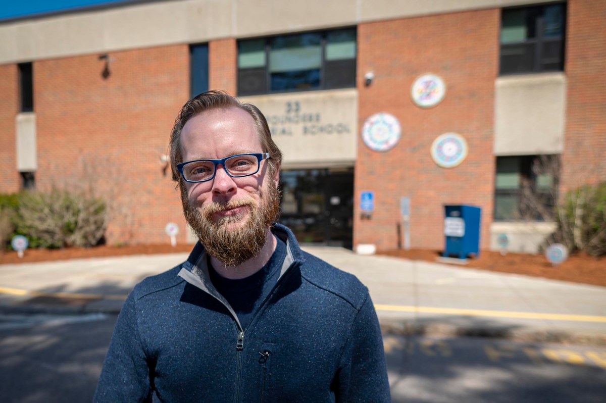 A man with glasses and a beard stands outside a brick school building on a sunny day.