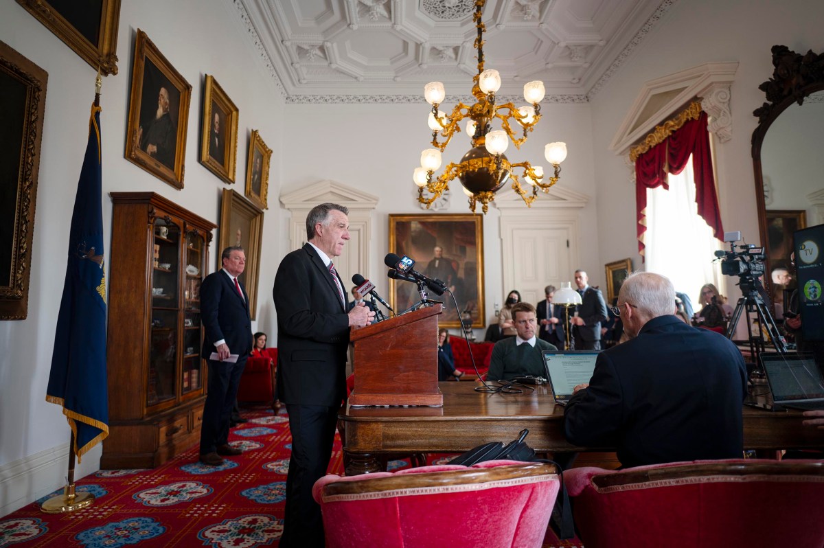 A man speaking at a wooden podium in an ornate room with chandeliers, surrounded by seated and standing attendees. Large portrait paintings adorn the walls.