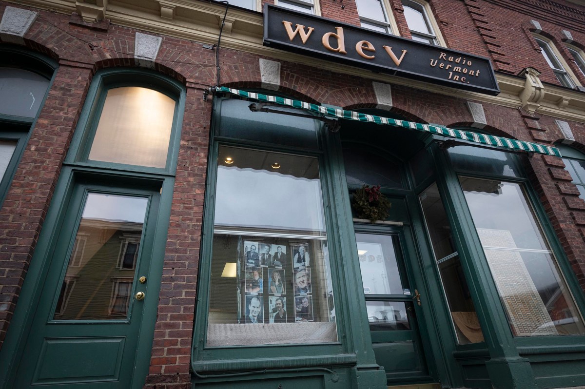 Exterior of WDEV Radio Vermont Inc. building with a green awning and large windows displaying photos. Brick facade under a sign with the station's name.
