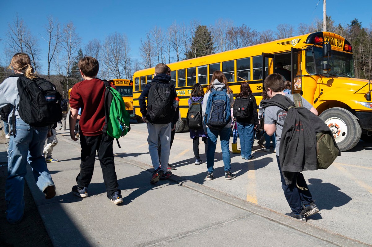 Students with backpacks walk toward a yellow school bus parked on a sunny day, with leafless trees visible in the background.