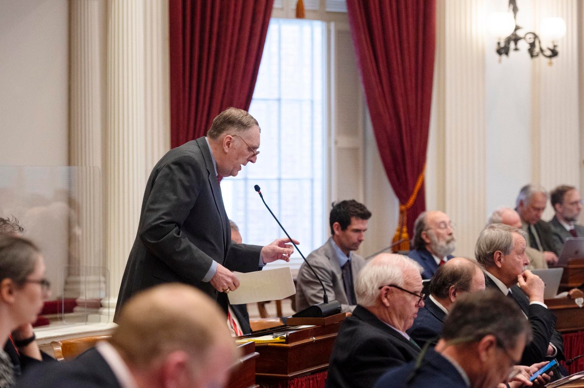 A man in a suit speaks at a podium in a formal meeting room with red curtains, surrounded by seated people, some using laptops.