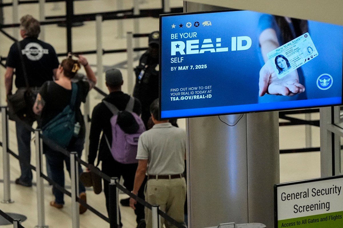 People wait in line at an airport security checkpoint near a sign promoting the requirement for REAL ID by May 7, 2025.
