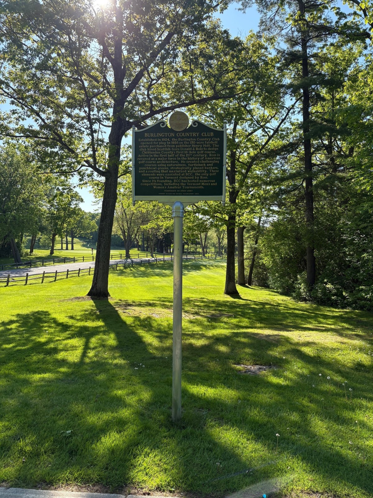 A historical marker for Burlington Country Club stands on a grassy area surrounded by trees, with a road and wooden fence visible on the left.