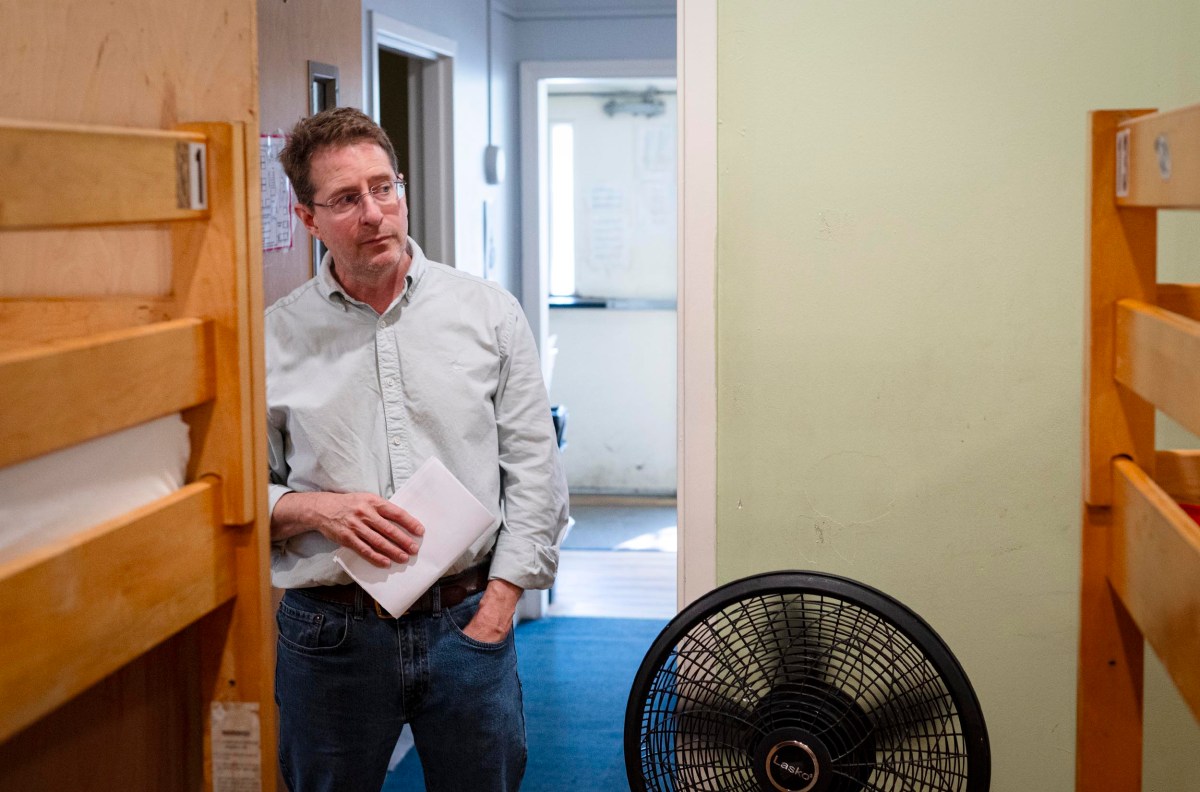 A man in glasses and a light shirt stands in a room between two bunk beds, holding papers, with a fan on the floor nearby.