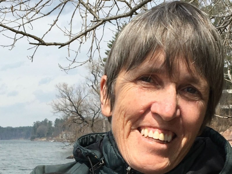 A person with short hair and a green jacket smiles outdoors near a lake, with bare trees and a cloudy sky in the background.