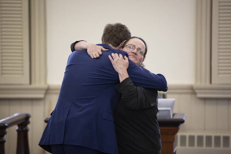 A man in a blue suit hugs a woman in black glasses and a black top inside a courtroom, with benches and windows in the background.