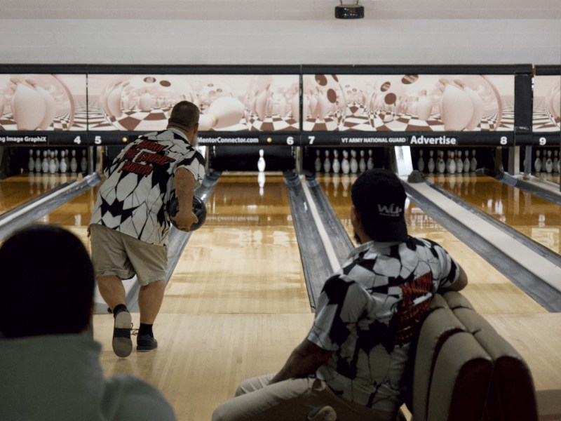 A man bowls while another team member watches from the seating area at a bowling alley with multiple lanes and pins set up.