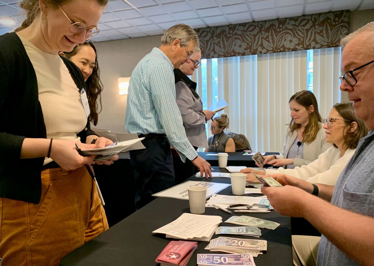 People participate in a tabletop activity involving paper money and documents at a conference or workshop setting, with several people seated and others standing.