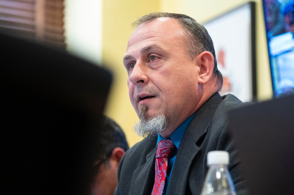 A man in a suit and red tie speaks at a meeting, with a water bottle and blurred objects in the foreground.