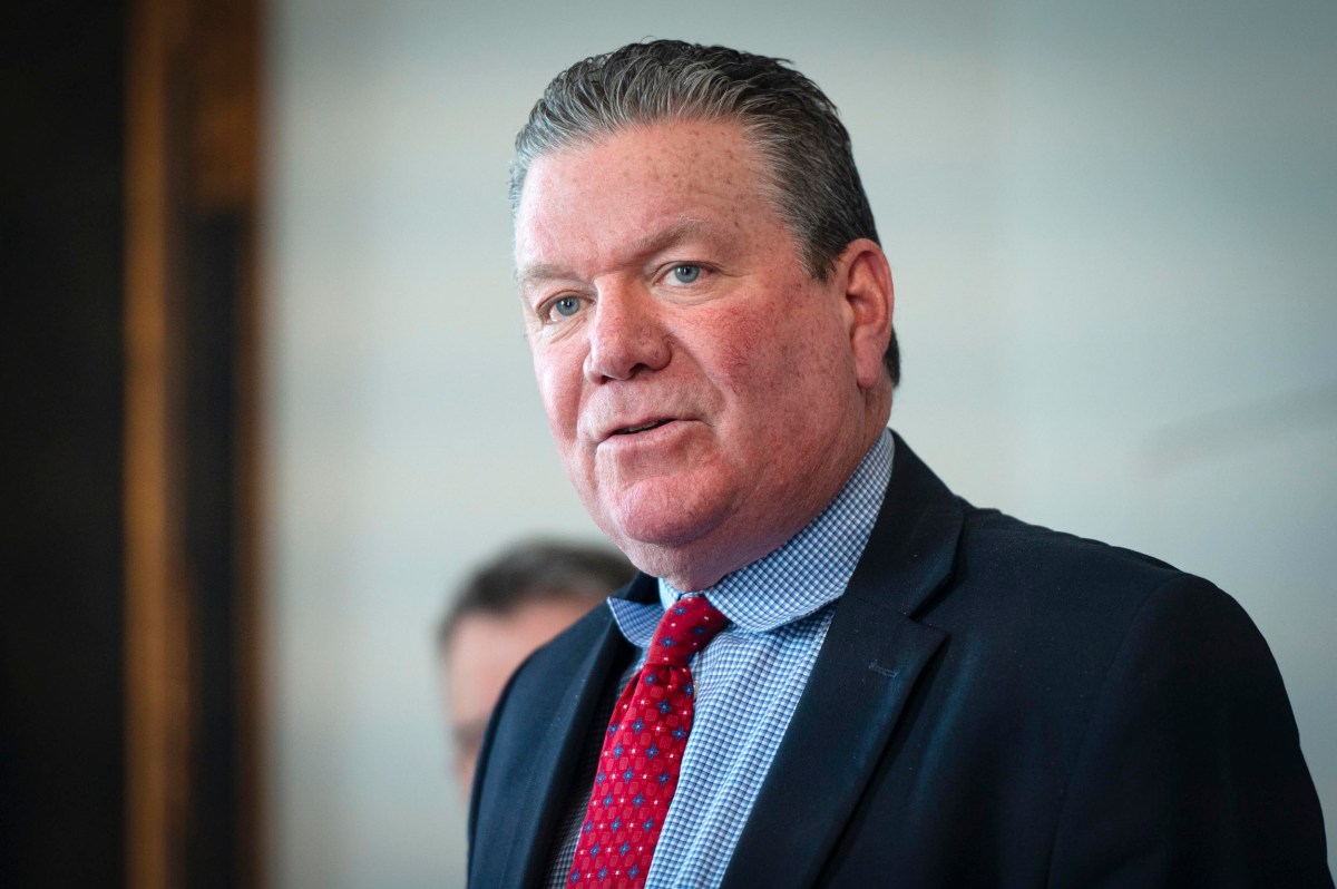 A middle-aged man in a suit and red tie speaks at an indoor event, facing the camera with a neutral background.