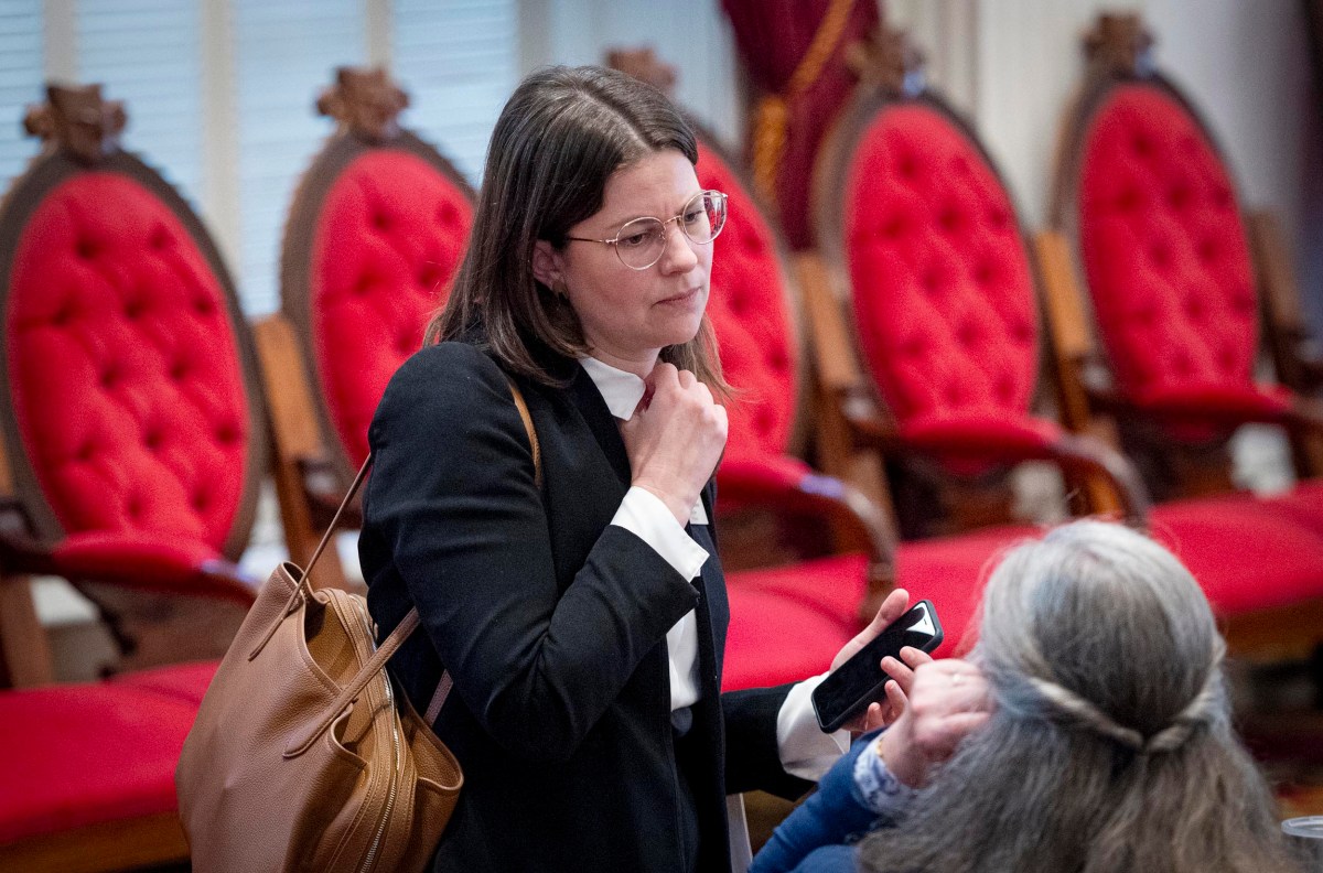 A woman in glasses and a black blazer stands holding her phone and speaking with another person in a room with red cushioned chairs.