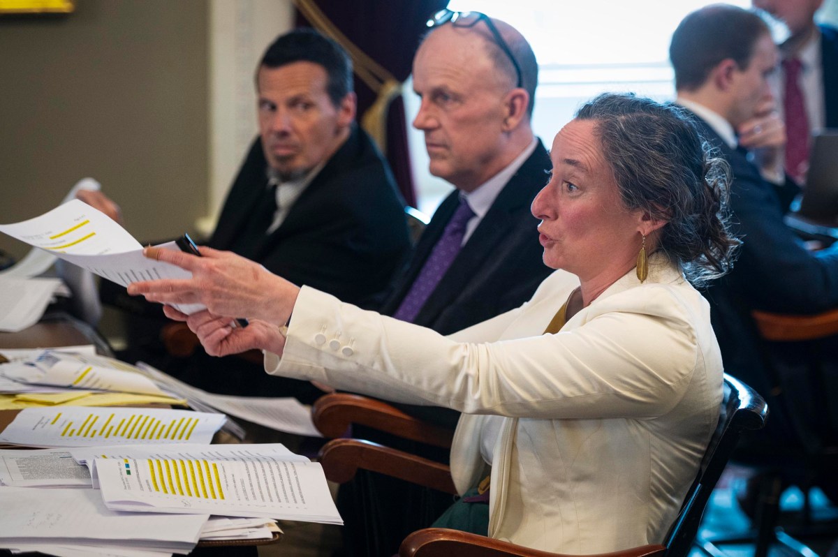 A woman in a white jacket gestures while holding papers at a meeting table with two men, surrounded by documents and other attendees in the background.