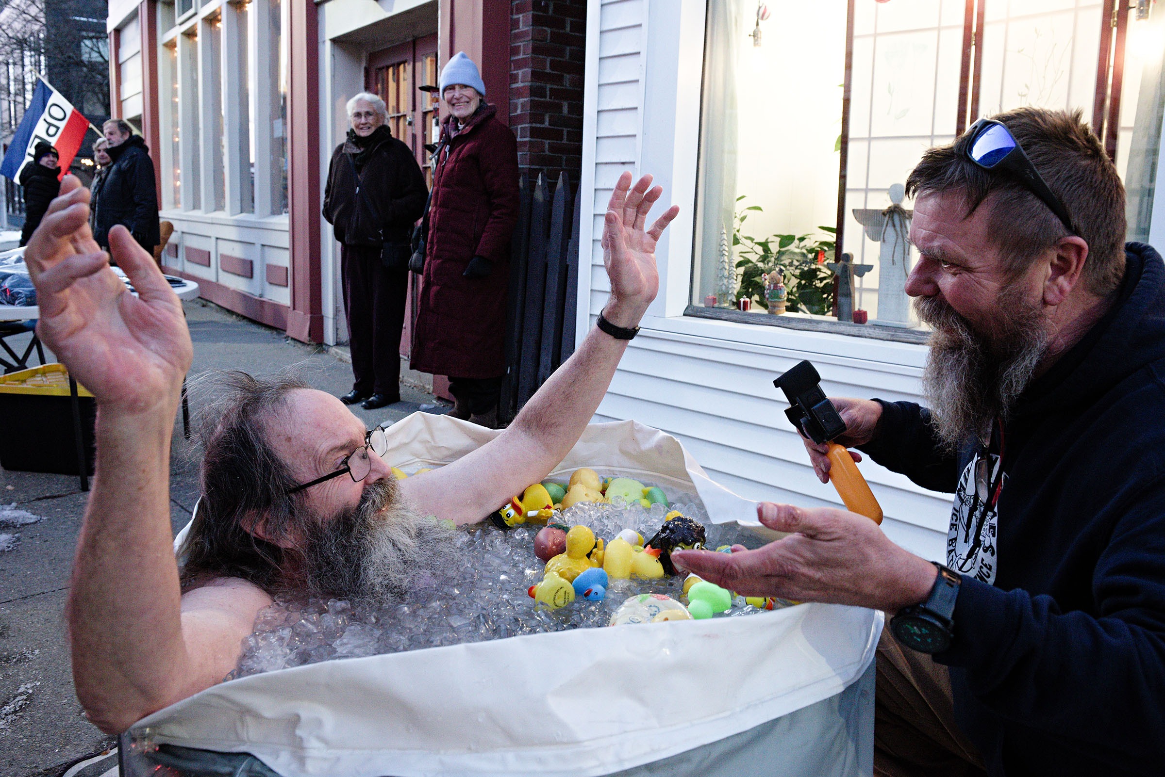 A bearded man sits in a tub filled with ice and rubber ducks on a sidewalk, raising his arms, while another man beside him holds a timer. Onlookers stand nearby and watch.