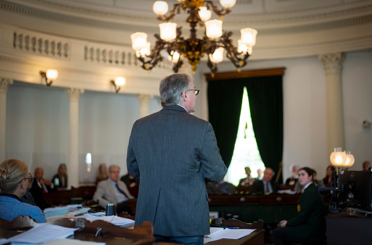 A man in a suit speaks to a seated audience in a formal, ornate room with chandeliers and columns.