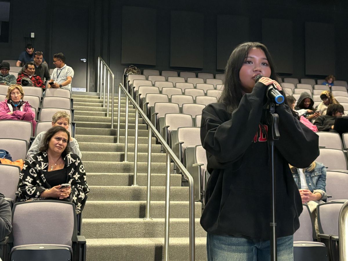 A young woman stands at a microphone speaking in an auditorium, while people sit and listen in the background.