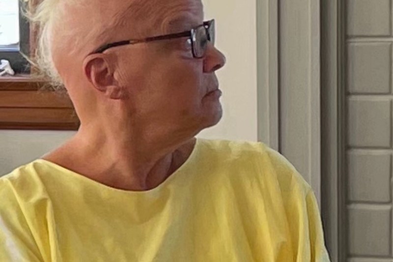 A person with short, light-colored hair and glasses wearing a yellow shirt is looking to the side indoors.