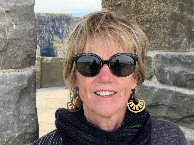 A woman with short hair and sunglasses smiles in front of a stone wall with cliffs visible in the background. She wears large earrings and a dark scarf.