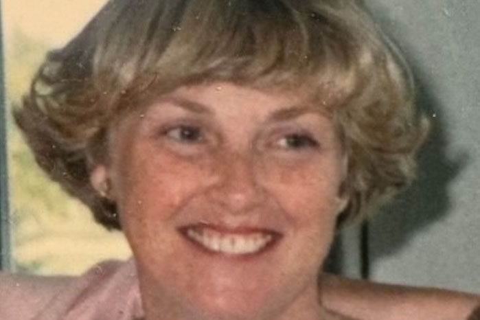 A woman with short, light-colored hair smiles while looking slightly to the side. She is wearing a light-colored top.