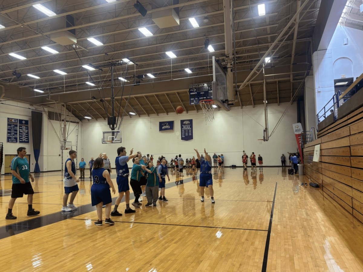A group of players in blue and teal jerseys play a basketball game in a gymnasium, with one player shooting the ball toward the hoop as spectators watch from the bleachers.