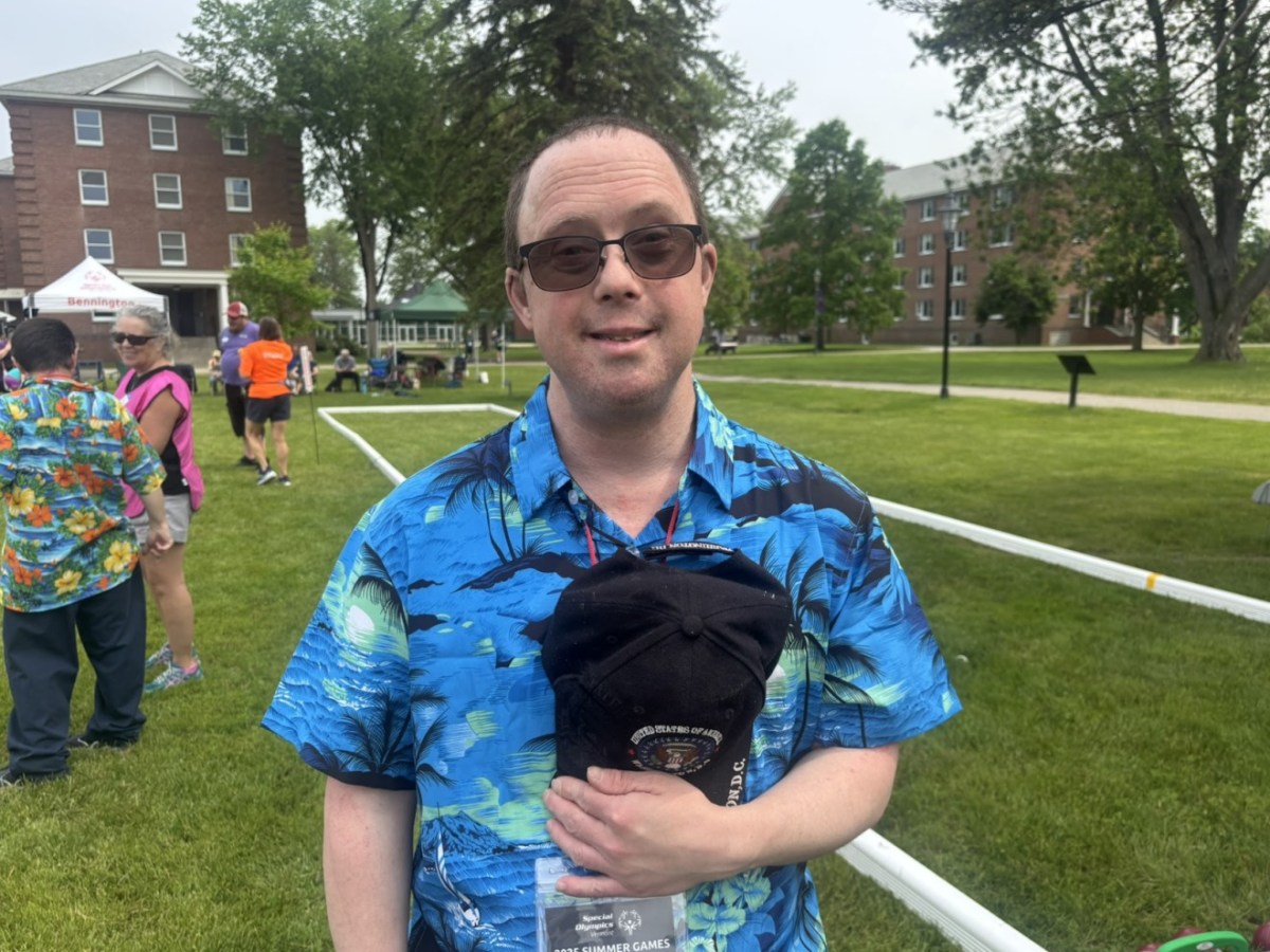 A man wearing sunglasses and a blue Hawaiian shirt stands on a grassy field, holding a black cap. People and buildings are visible in the background.