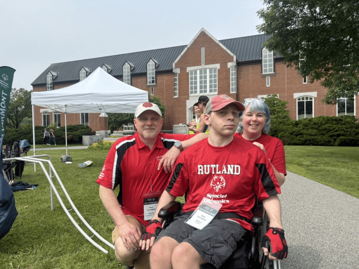 Three people, two men and one woman, wearing red "Rutland" shirts, pose outside a brick building with lawn and white tent in the background.