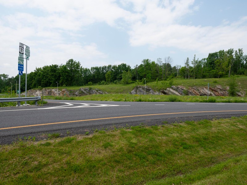 A rural road intersection with route signs, a guardrail, and grassy areas alongside tree-lined rocky terrain under a partly cloudy sky.