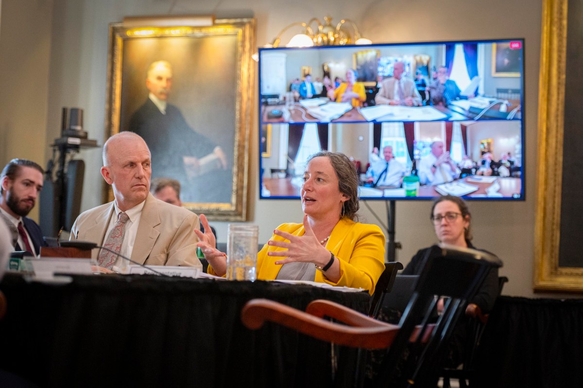 A woman speaks at a conference table with others, while a man listens; multiple participants appear on a large screen in the background during a meeting.