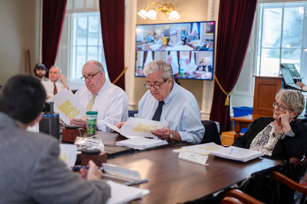 Several people sit at a conference table reviewing documents, with video screens and remote participants visible in the background.