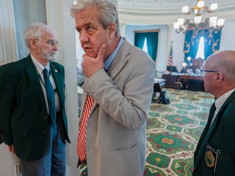 Three older men stand in a formal room with ornate carpet; one man in a suit touches his chin while two others in green blazers watch.