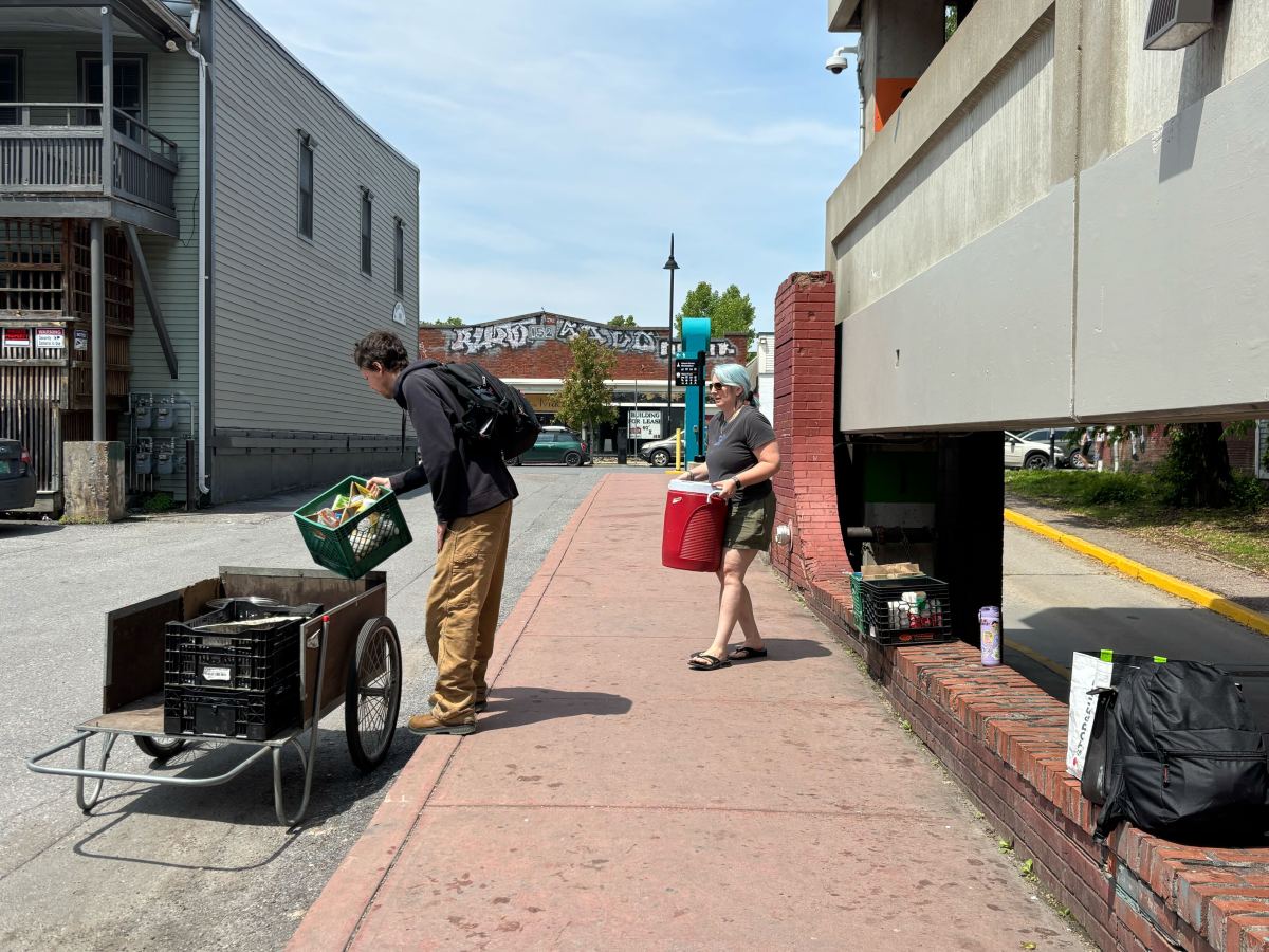 Two people load supplies into a cart on a sidewalk next to a parking garage; one holds a crate, the other carries a red cooler.