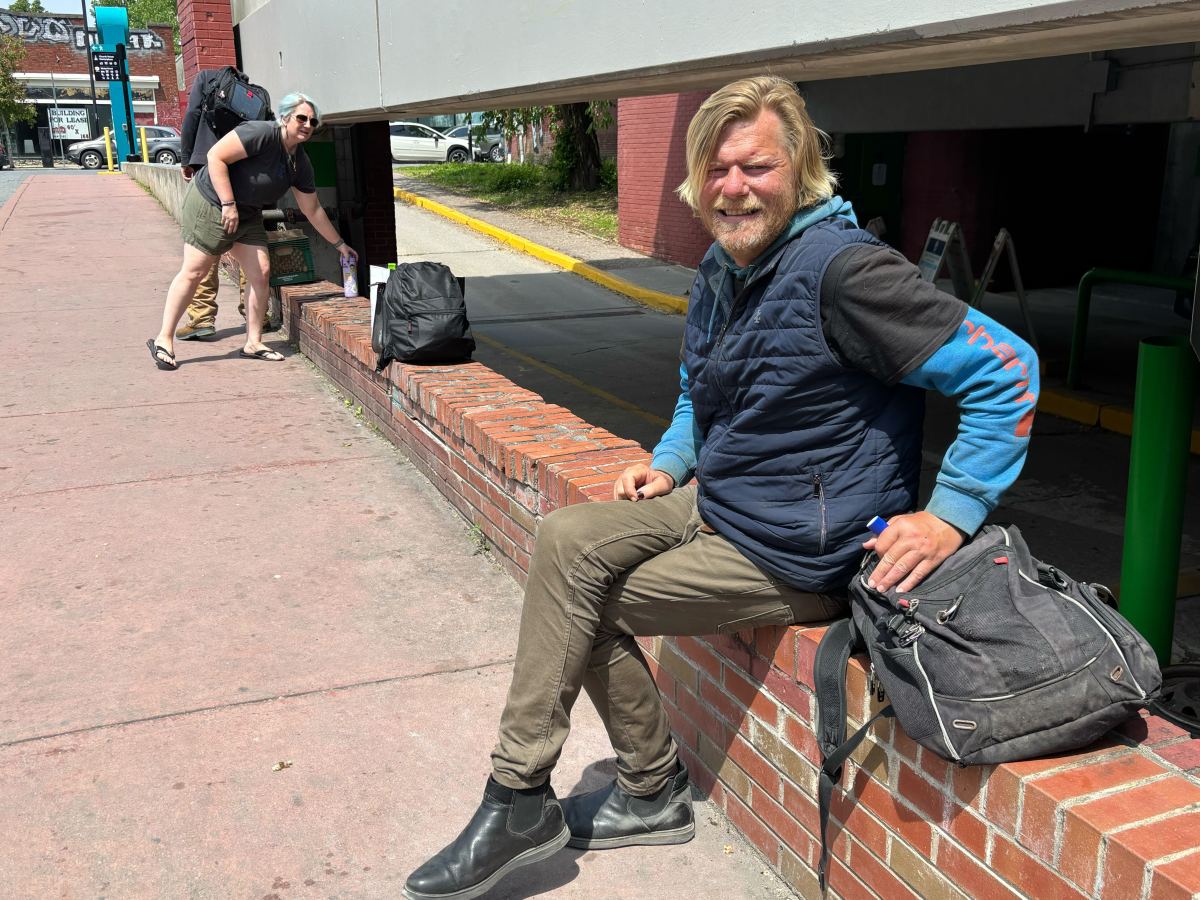Two men with backpacks are outside; one is sitting on a brick ledge smiling at the camera, while the other is standing in the background adjusting his bag.