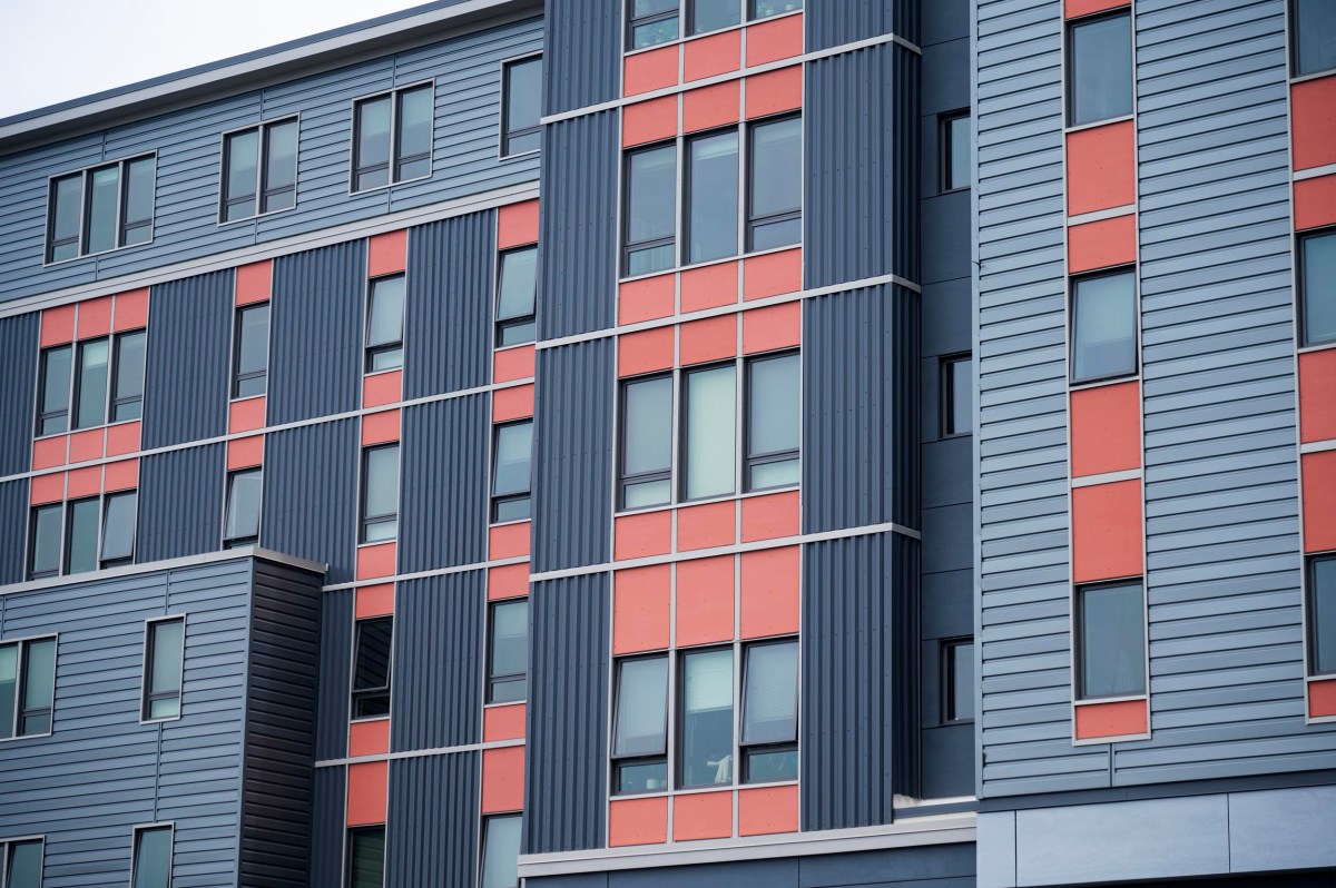 A close-up view of a modern apartment building facade with gray and red panels, vertical and horizontal lines, and multiple windows.