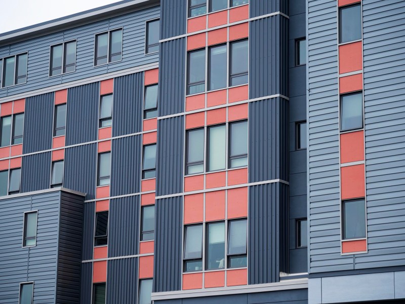 A close-up view of a modern apartment building facade with gray and red panels, vertical and horizontal lines, and multiple windows.