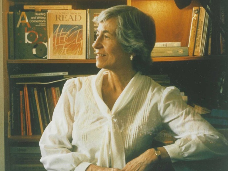A woman with a white blouse sits in front of a bookshelf filled with books, looking to the side and smiling.
