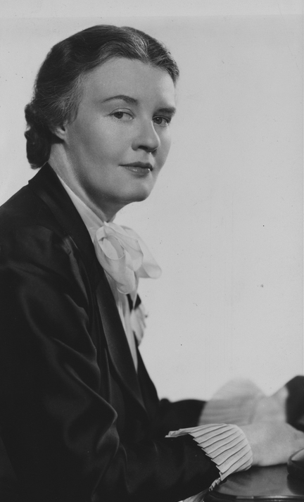 Black and white portrait of a woman with dark hair, wearing a suit jacket, a pleated blouse, and a large bow at the neck, looking slightly to the side.