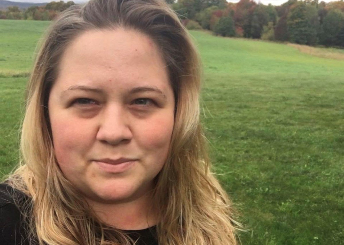 A woman with long, blonde hair stands outdoors in front of a green field with trees and a cloudy sky in the background.