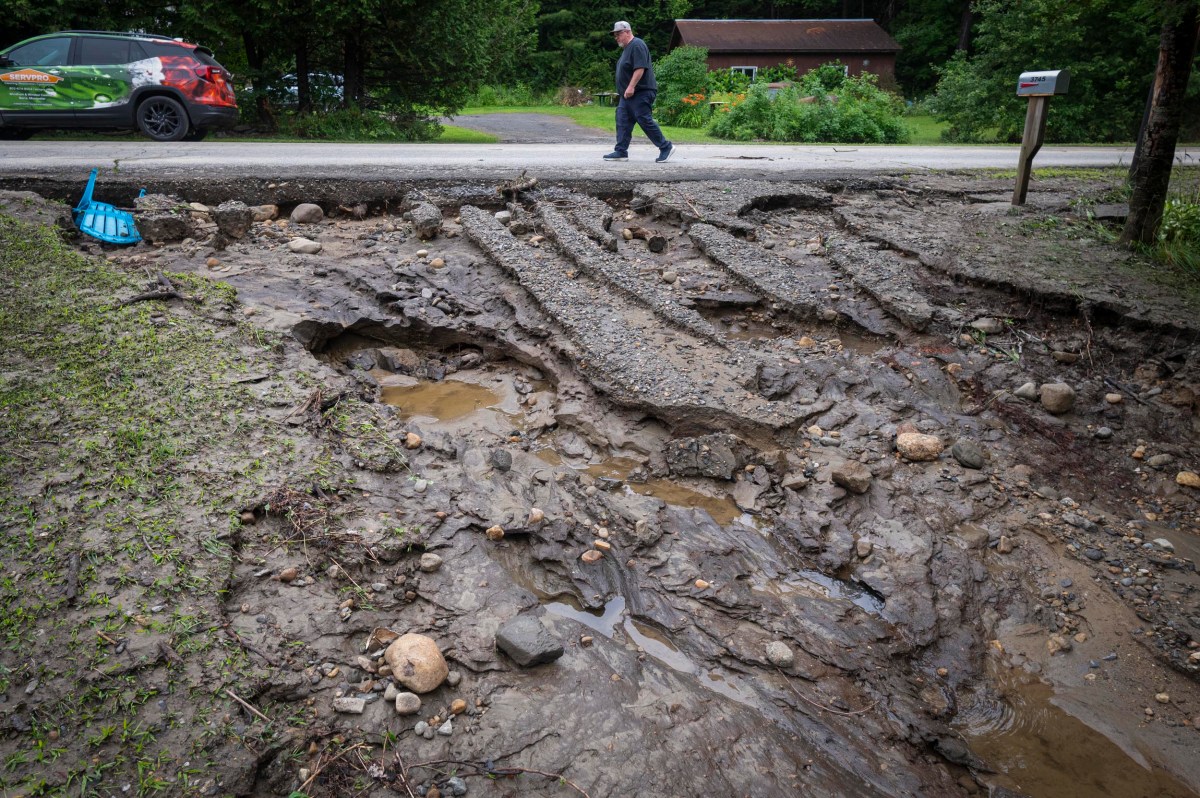 A man walks past a road damaged by erosion, with large sections of asphalt and soil washed away, exposing rocks and muddy water.