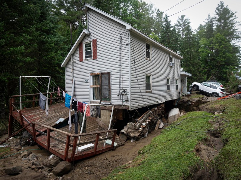 A white house with a collapsed porch and damaged foundation leans over an eroded slope; clothes hang on a line and a car is partially submerged in mud nearby.
