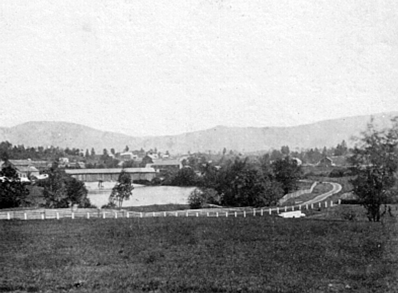 Black and white photo of a rural landscape with a pond, trees, fenced fields, and distant buildings set against rolling hills.