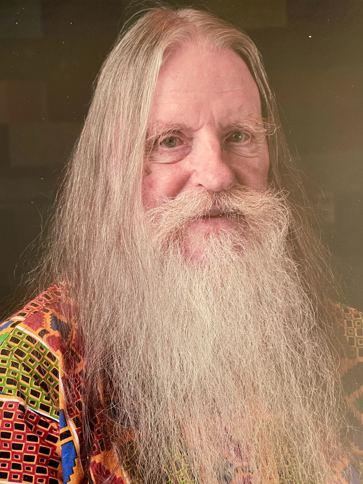 A man with long hair and a full beard, wearing a colorful geometric-patterned shirt, poses against a dark background.