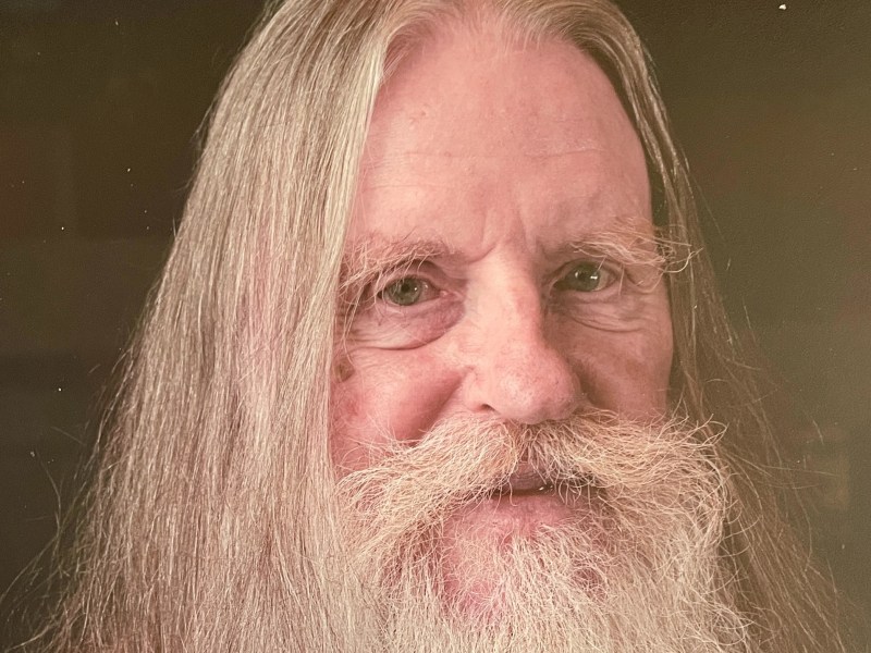 A man with long hair and a full beard and mustache looks directly at the camera, against a dark background.