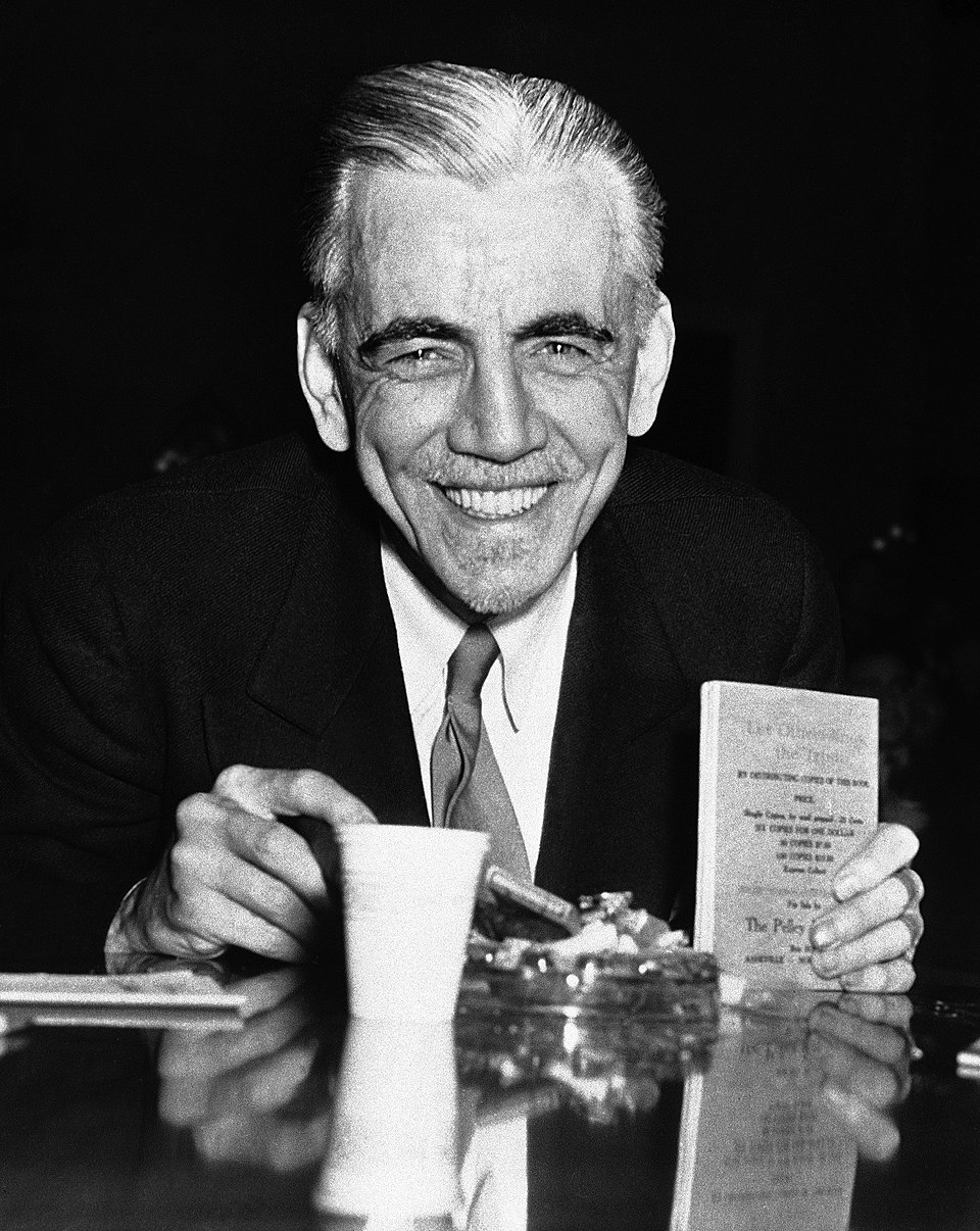 A smiling man in a suit sits at a table holding a booklet, with a cup and an ashtray in front of him.