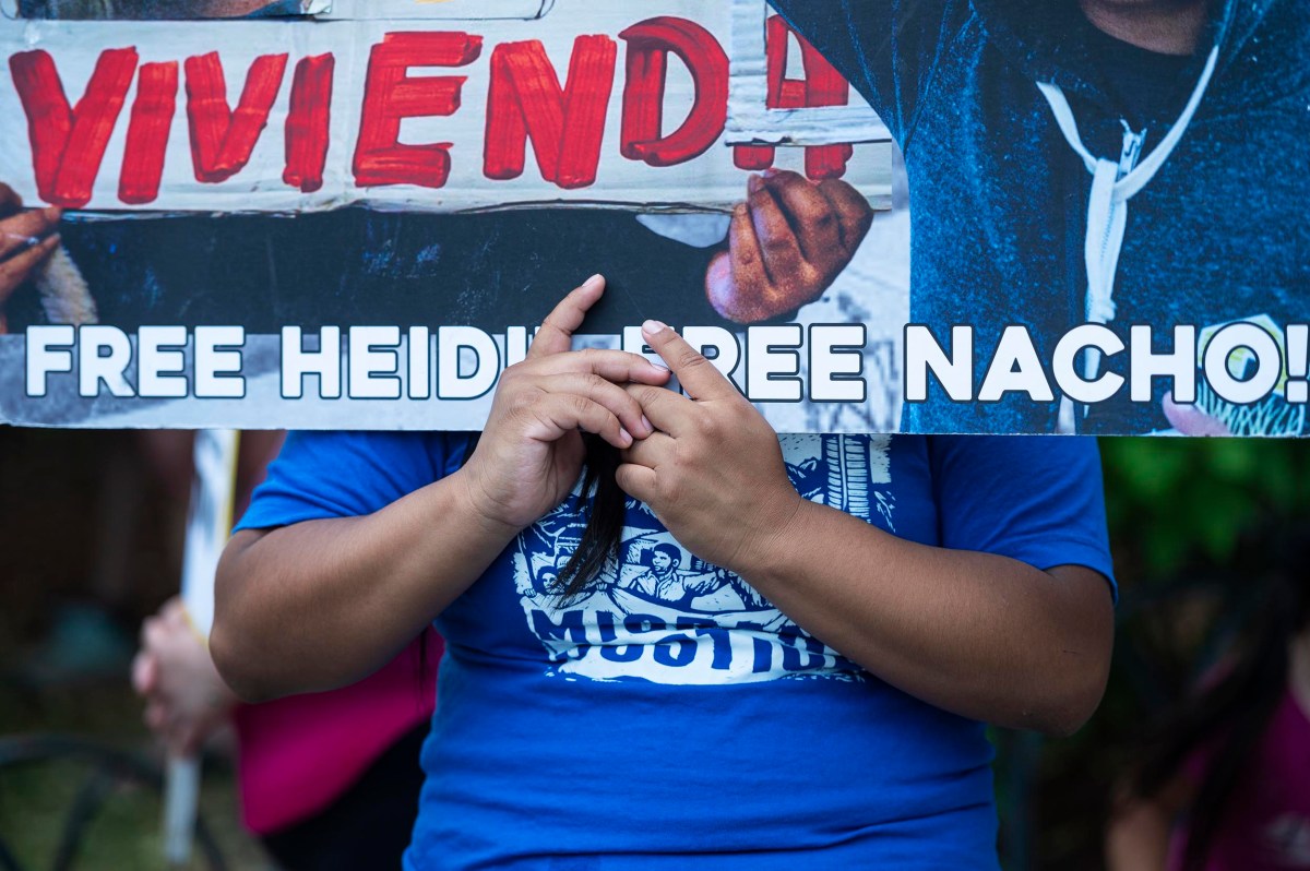 A person holds a banner that reads "FREE HEIDI FREE NACHO!" and part of the word "VIVIENDA," with their hands visible in front of their torso.