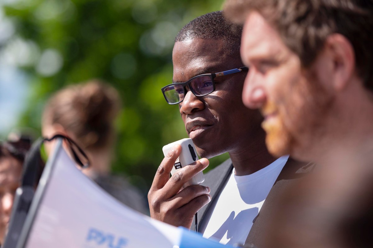 A man holds a microphone and speaks at an outdoor event, surrounded by other people, with greenery in the background.