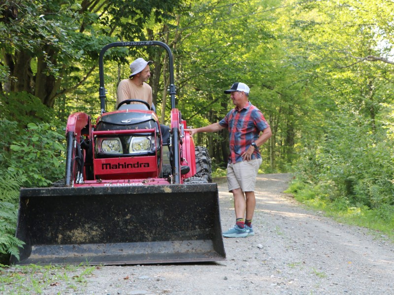 Two men talk on a dirt road; one sits on a red Mahindra tractor while the other stands beside it amidst a green, wooded area.